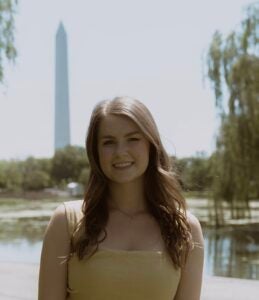 Hannah is a younger white woman with long, light brown hair. Hannah is wearing a yellow shirt and standing in front of the Washington Monument. 