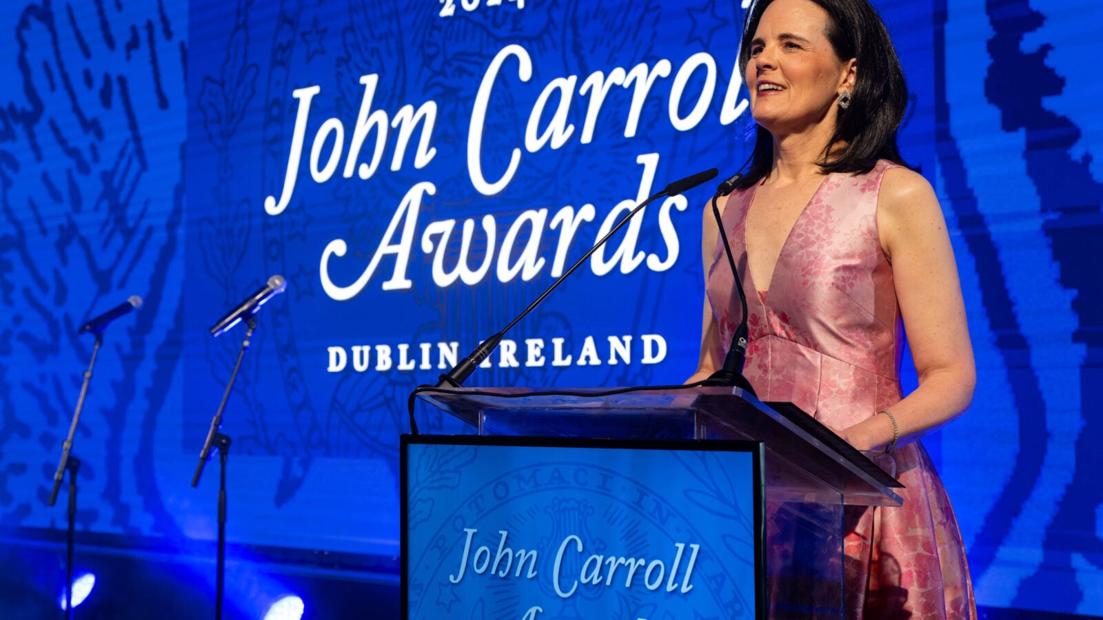 a woman in a pink dress stands at a podium in front of a screen with the words 