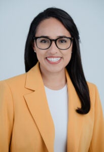 a woman with dark hair and glasses wears an orange blazer and a white shirt