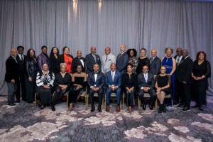 A diverse group of 25 people, dressed in formal attire, poses in two rows against a gray curtain. The atmosphere is celebratory and professional.