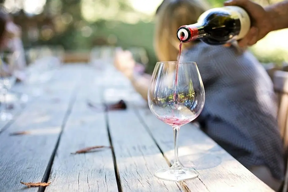 A person pours a red wine into a wine glass on a wooden picnic table.